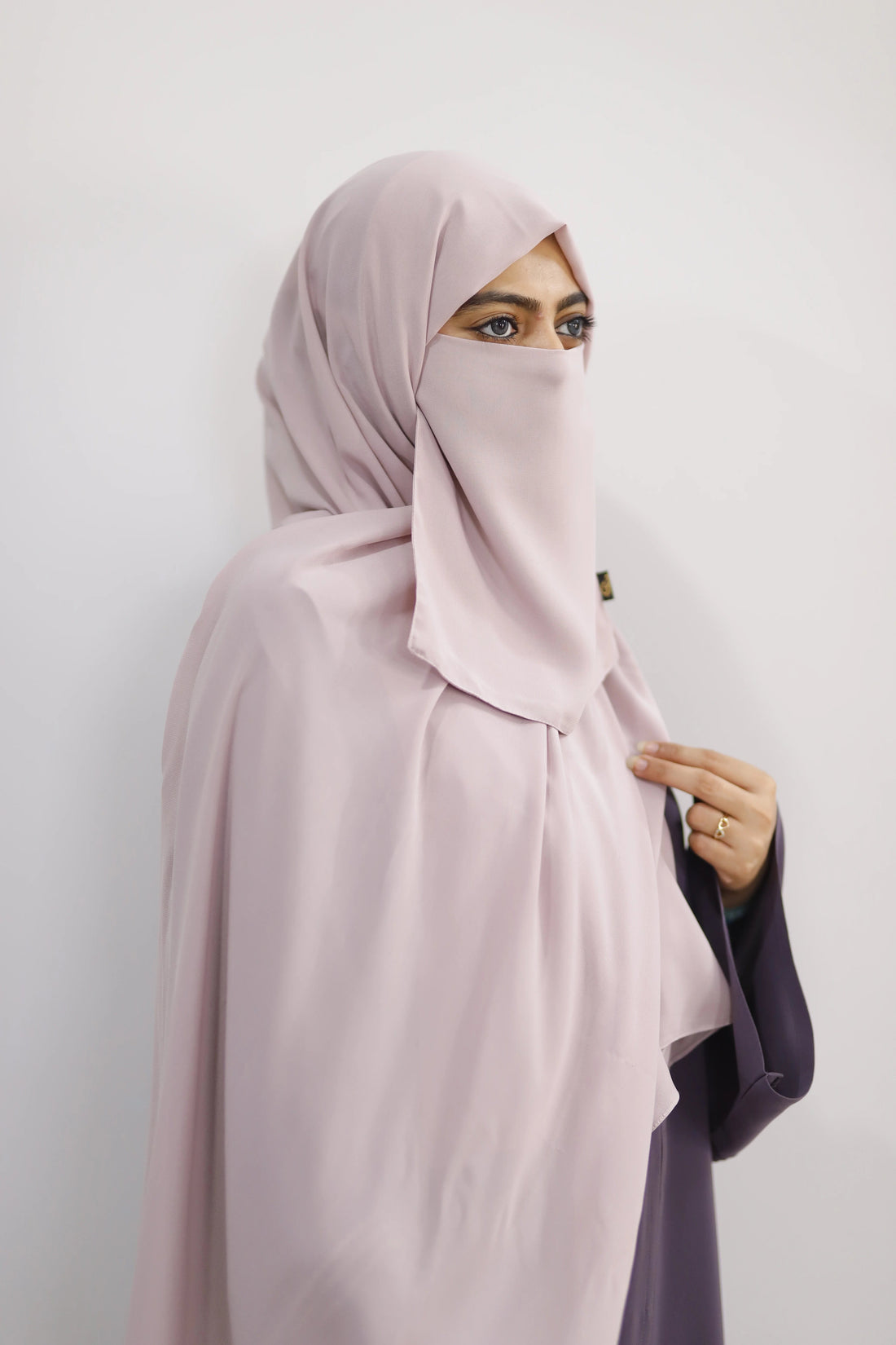 Person wearing a light pink hijab and purple garment on a white background
